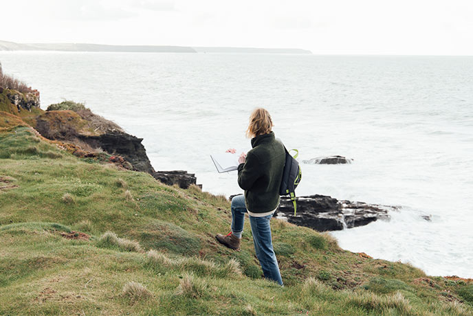Emma Scattergood walking with her sketchbook on some Cornish cliffs above the sea