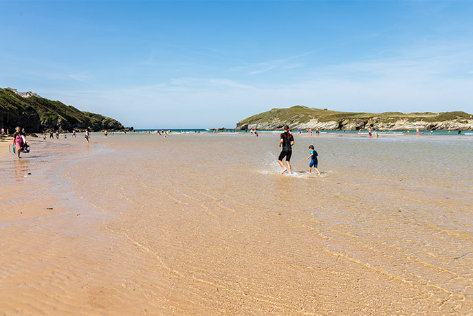 A father and child running through the shallow water on the golden sandy beach at Porth, one of the best beaches near Newquay