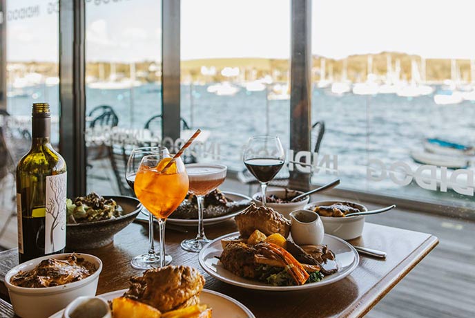 A table full of food and cocktails with sea views in the background at INDIDOG in Falmouth