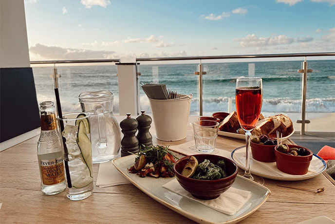A selection of dishes and drinks on a table at Porthmeor Beach Café with sea views in the background