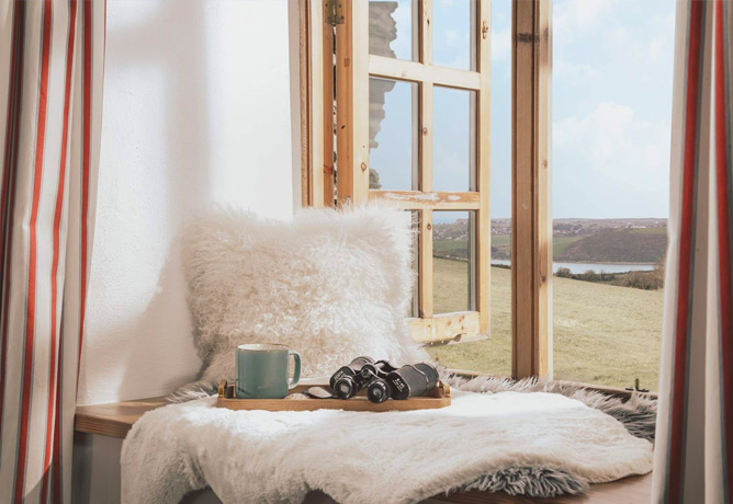 A window seat complete with pillow, mug, book and binoculars looks out over the fields and down to the water