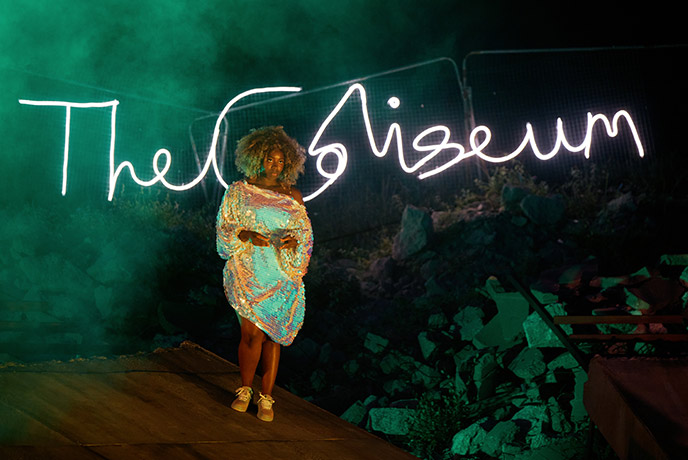 An actress portraying Scylla wearing a sequined dress and standing in front of a neon Coliseum sign during a Wildworks production of I Am Kevin