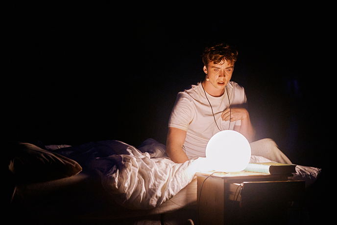 An actor sitting on a bed in the dark staring at a lamp in the shape of the moon