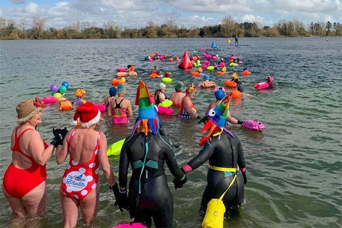 A group of people wearing swimming costumes and Santa hats swimming in a like in the Cotswolds at Christmas