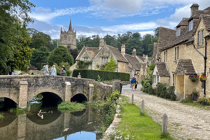An ancient bridge, babbling brook and honey hued cottages with a church in the background in Castle Combe