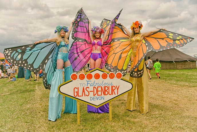 Women wearing stilts and costumes with the Glas-denbury sign
