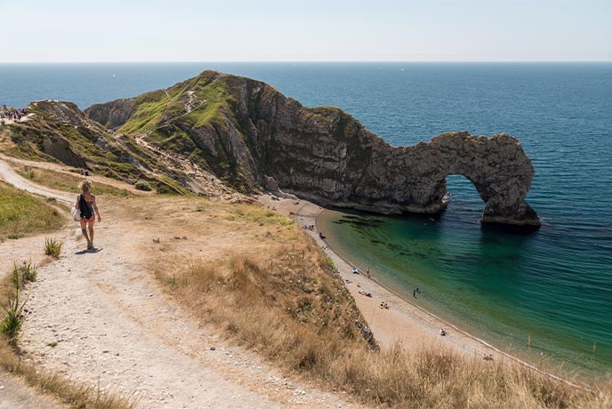 Someone walking down to the golden beach at Durdle Door, with a stone arch in the background