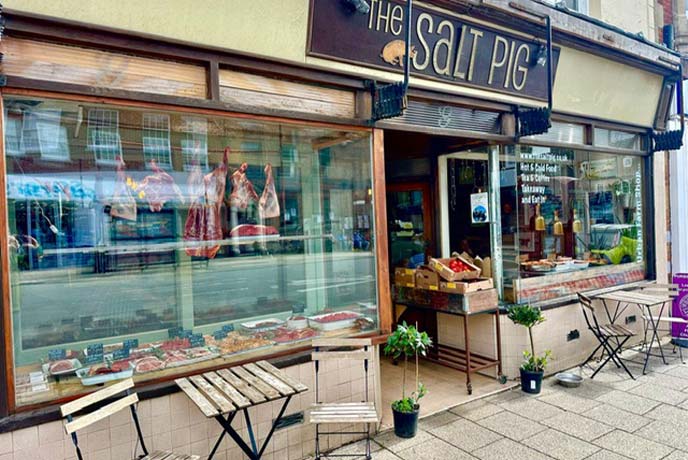 The charming store front of The Salt Pig farm shop in Dorset with wooden tables and chairs out front and meat in the windows