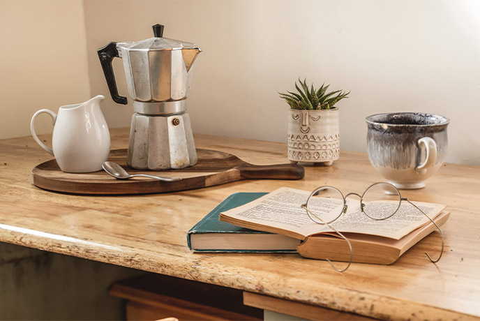 A book and a pair of reading glasses on a table with a coffee pot in the background