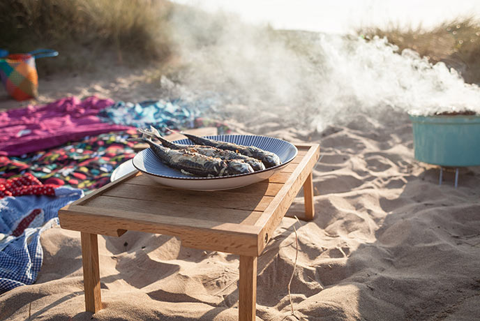A lit BBQ and cooked mackerel on the beach surrounded by towels