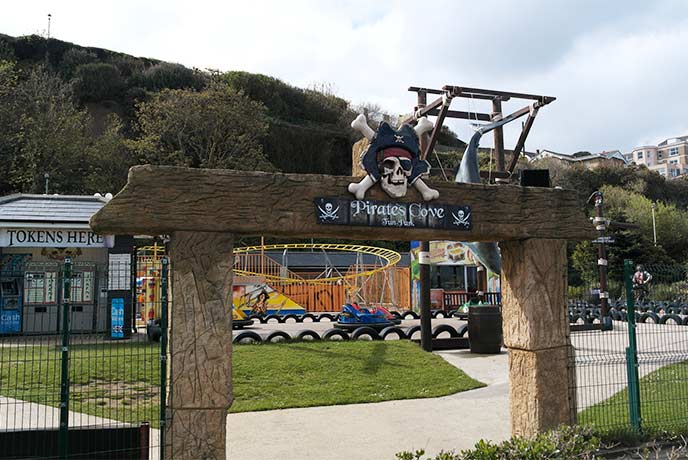 A large skull and crossbones above the entrance to Pirates Cove Crazy Golf in Shanklin with rides in the background