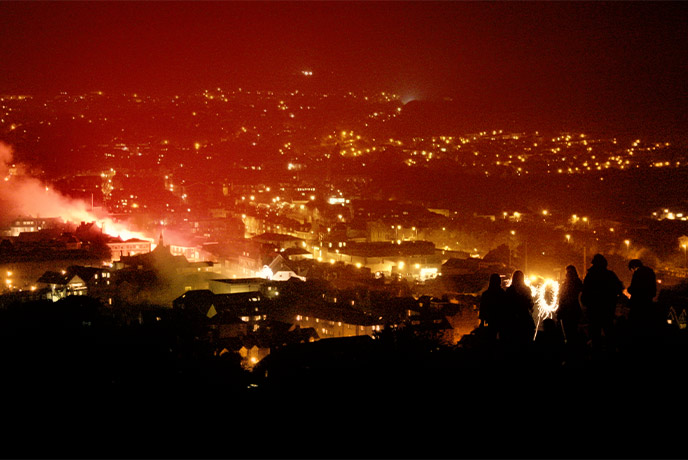 The silhouettes of people overlooking Lewes during Lewes Bonfire Night, with lots of red smoke coming out of the town