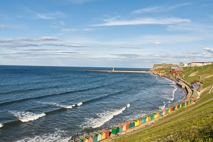 Looking down over grassy cliffs at colourful beach huts and the sea at West Cliff beach in Whitby
